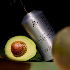 Acure body lotion next to an avocado on a dark background