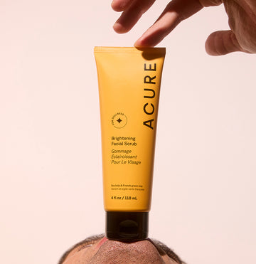 Man holding a tube of Acure Brightening Facial scrub against his chin, holding the top with his hand