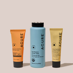 Three ACURE brand travel essential products including a sunscreen tube, a dry shampoo, and a 12-hour moisturizer, each in their distinctive packaging.