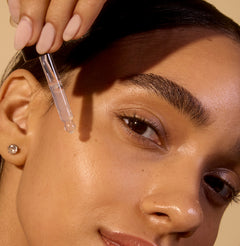 Close-up of a woman applying a dropper of liquid to her eyebrow against a beige background