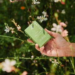 Hand holding Avocado Oil and Peppermint soap bar Acure x Apricot Lane Farm product box among flowers
