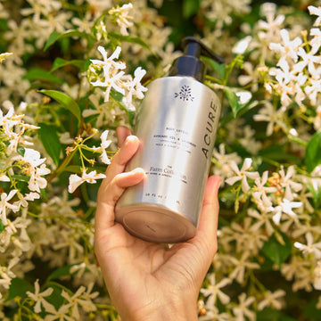Hand holding a silver bottle labeled 'ACURE Farm collection' against a floral background