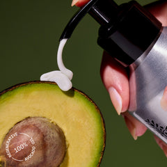 Hand holding a pump bottle with white cream being squeezed onto an avocado half against a green background