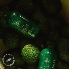 Two green bottles labeled 'ACURE x Apricot Lane Farm' on a bed of dark green spherical objects.