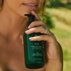 Person holding a green bottle labeled 'The Farm Collection' with a blurred natural background