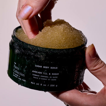Hand holding a jar of Acure Organics sugar body scrub with visible text on a plain background