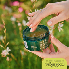 Person holding a green jar of body scrub with a blurred natural background