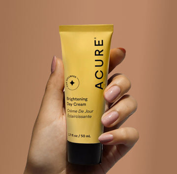 Hand holding a tube of Acure Brightening Day Cream against a brown background