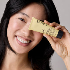 Woman holding a tube of Acure product near her face against a neutral background