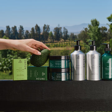 Acure skincare products on a table with a hand holding an avocado outdoors.