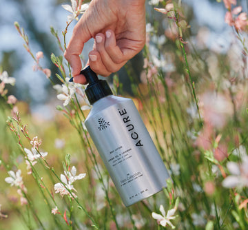 Hand holding Acure lotion bottle in a natural setting with flowers