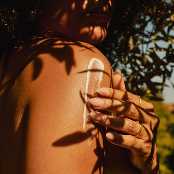 Person applying lotion to their shoulder with a palm leaf casting shadows.