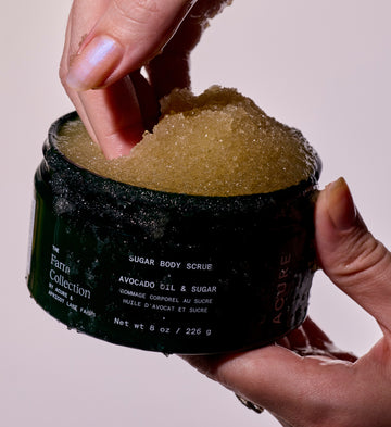 Woman taking Sugar body scrub from a dark jar labeled The Farm Collection by ACURE x APRICOT LANE FARMS