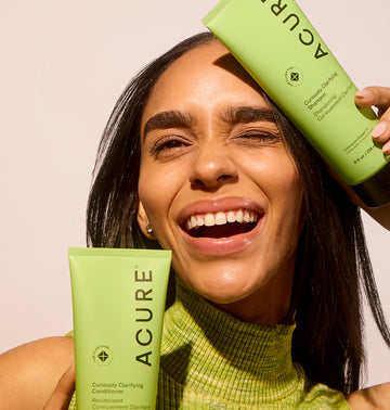 Woman holding Acure shampoo and conditioner bottles against a beige background